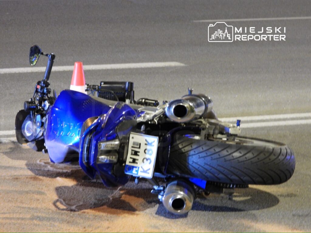 Nieoznakowany motocykl leży na boku na jezdni, z czerwonym pachołkiem obok.