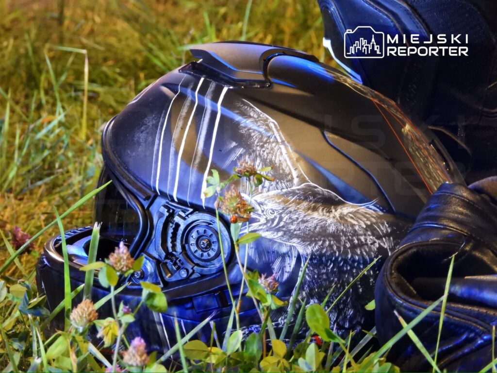 Czarny kask motocyklowy z grafiką orła leży na trawie, obok skórzana rękawica.