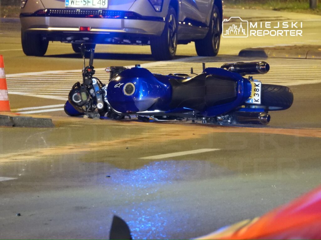 Nieprzewidziana sytuacja na drodze: przewrócony niebieski motocykl leży na jezdni obok pojazdu osobowego.