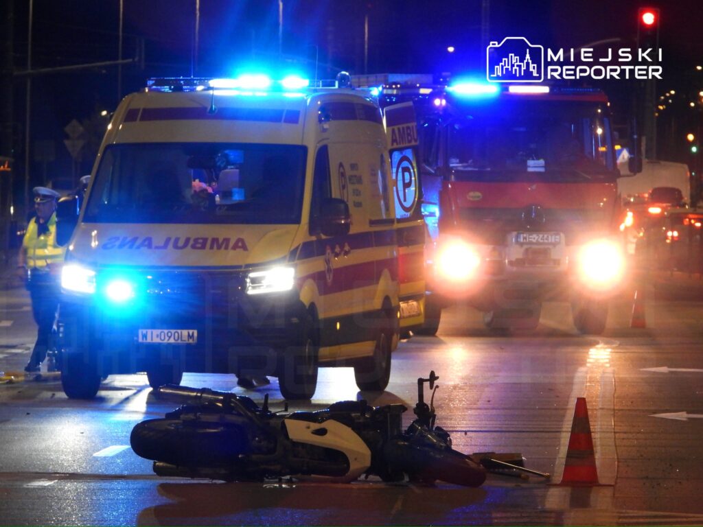 Karetka pogotowia z włączonymi sygnałami świetlnymi na drodze obok przewróconego motocykla, w tle woz strażacki.