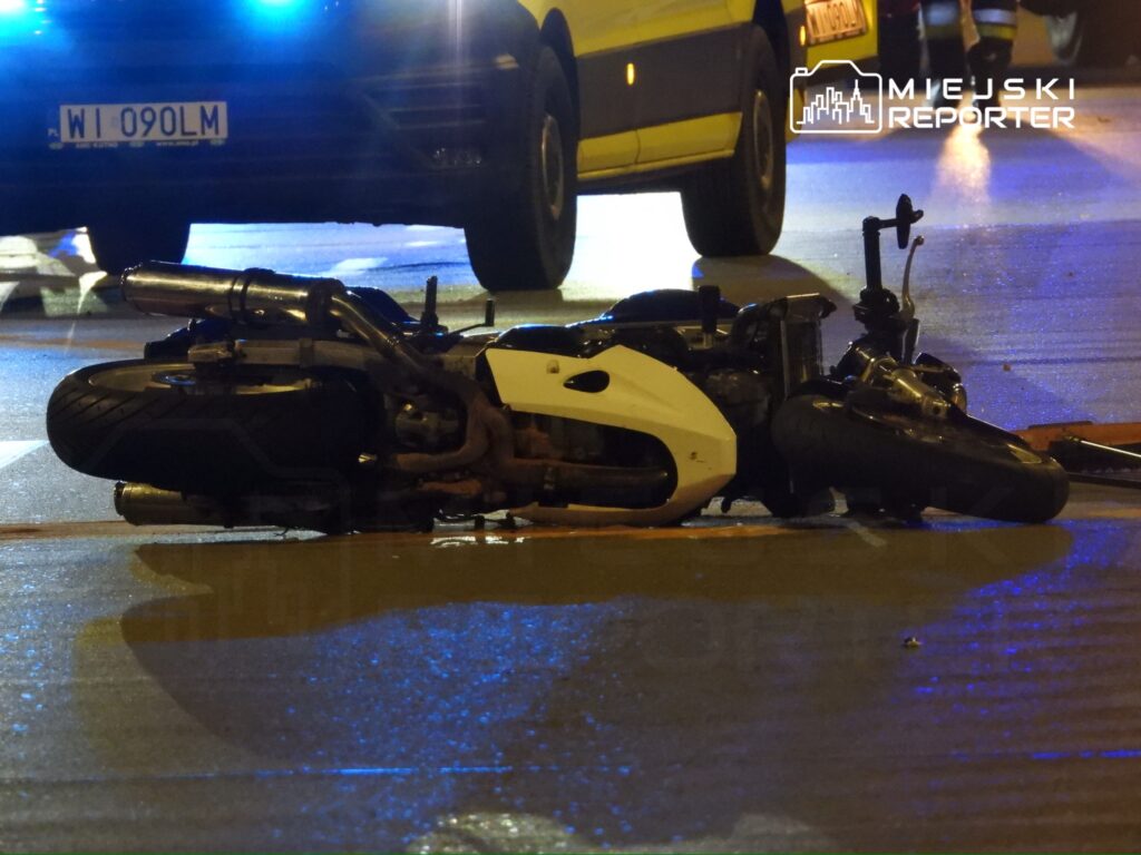 Motocykl leżący na jezdni, obok widoczny radiowóz policyjny z włączonymi sygnałami świetlnymi.
