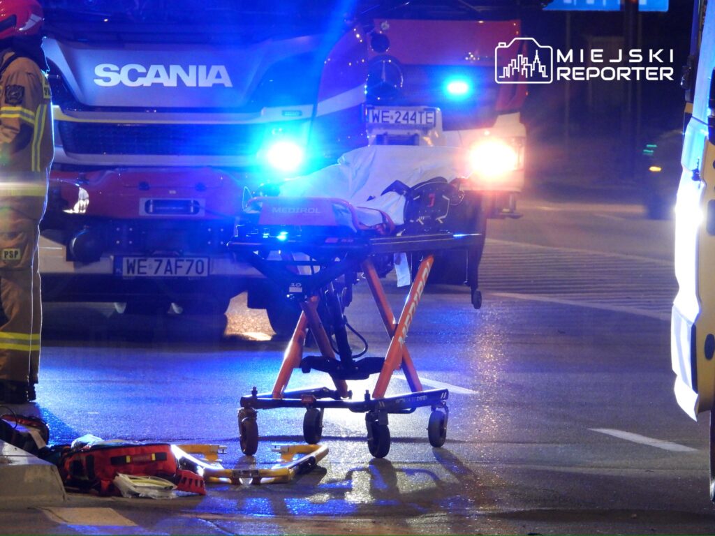 Strażak w mundurze stoi obok wózka medycznego na oświetlonej ulicy, w tle pojazdy ratunkowe.