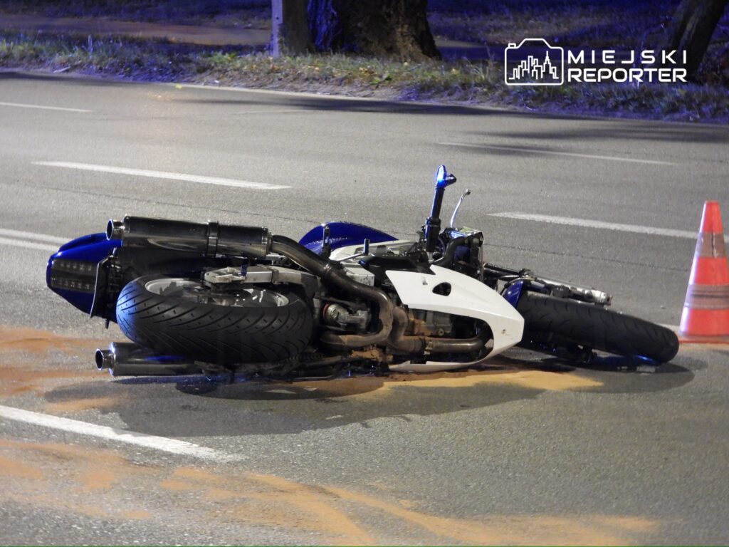Motocykl leży na jezdni, obok pomarańczowy trójkąt ostrzegawczy, a na asfalcie widoczna plama.