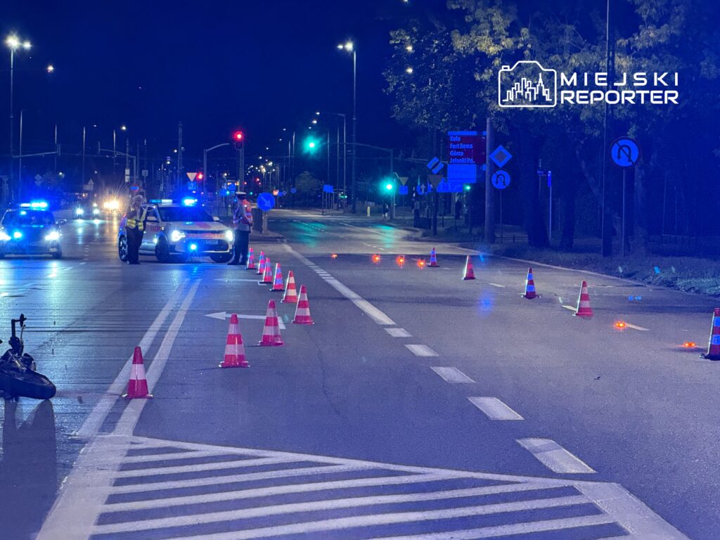 Policjanci z niebieskimi sygnałami kontrolują ruch na zamkniętej drodze, otoczonej pachołkami.