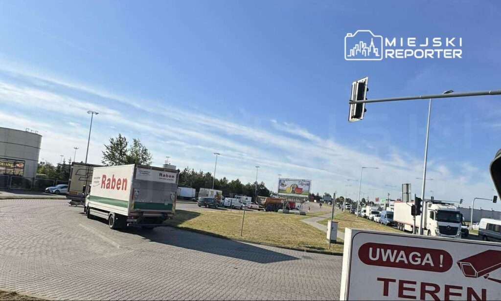 Ciężarówki marki Raben stoją w kolejce na rondzie, w tle widoczne reklamy i zieleń. Słoneczne niebo.