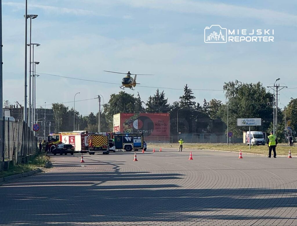 Helikopter ratunkowy unosi się nad terenem akcji, obok stoją pojazdy straży pożarnej i policji, a wokół wyznaczone strefy bezpieczeństwa.