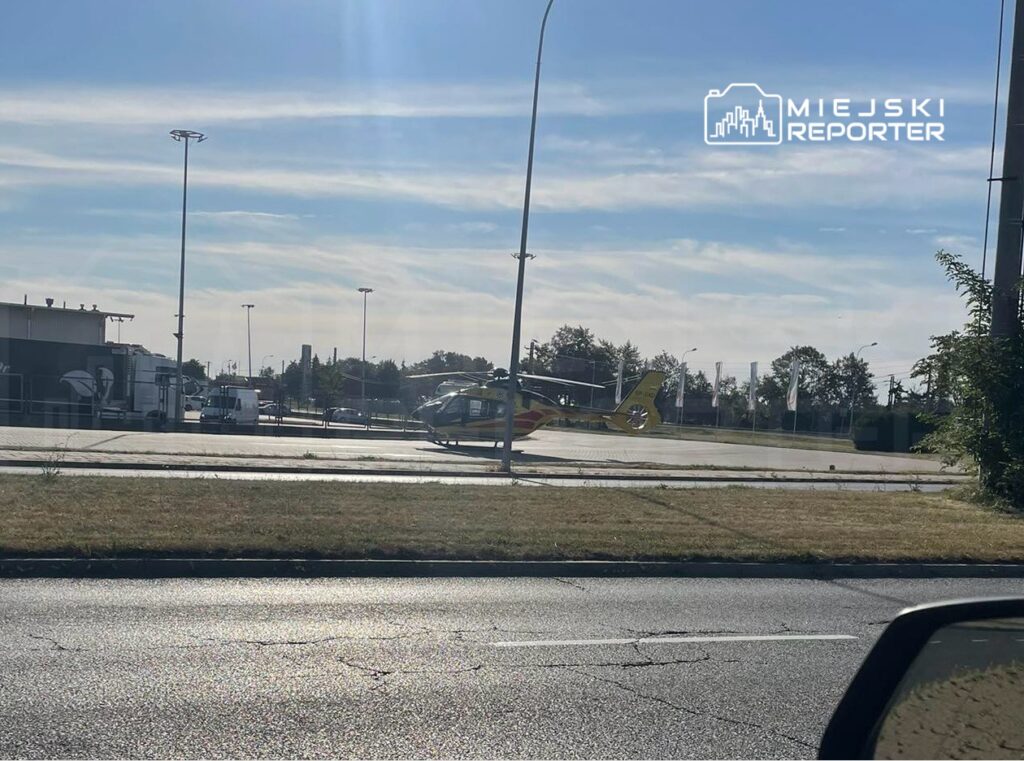 Helikopter lądował na parkingu w pobliżu budynków przemysłowych, otoczony przez pojazdy dostawcze.