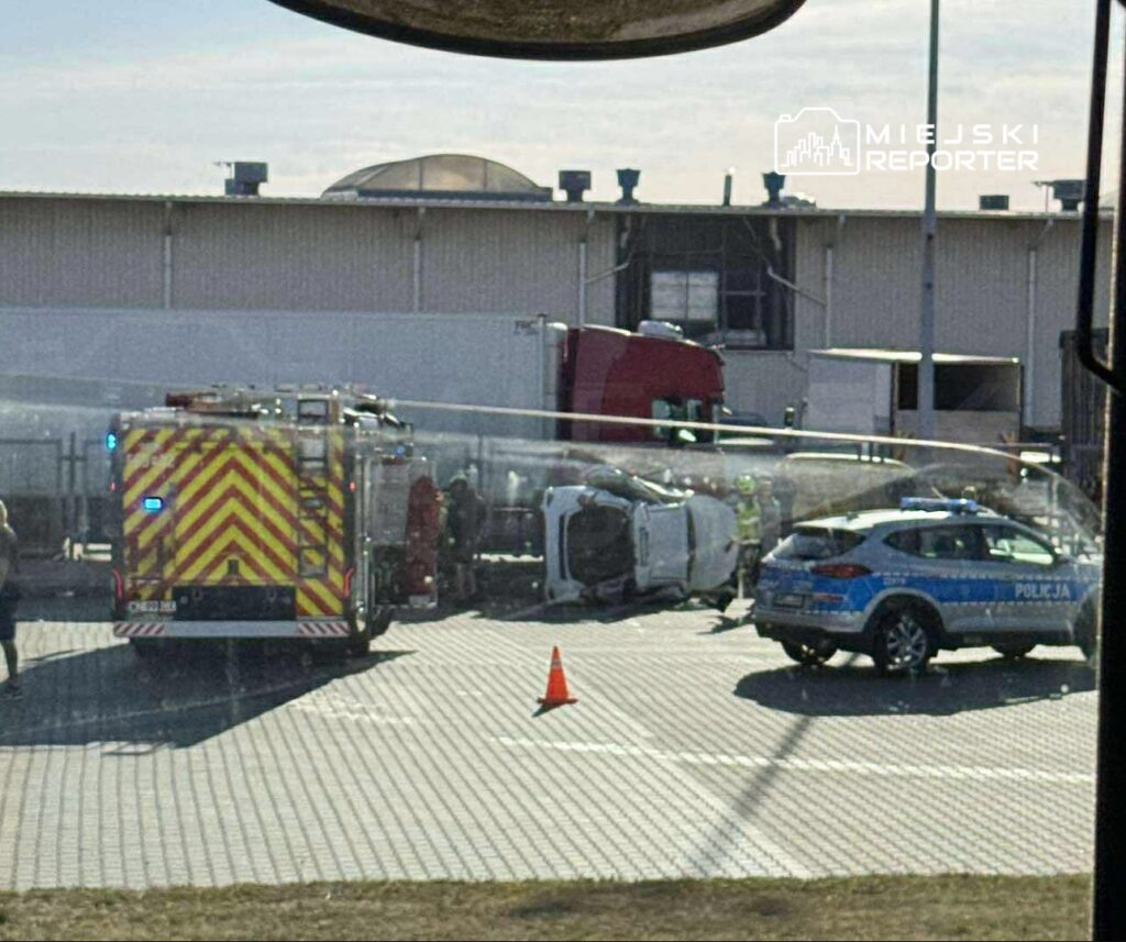 W miejscu wypadku samochodowego strażacy i policja pracują przy przewróconym pojeździe na parkingu.