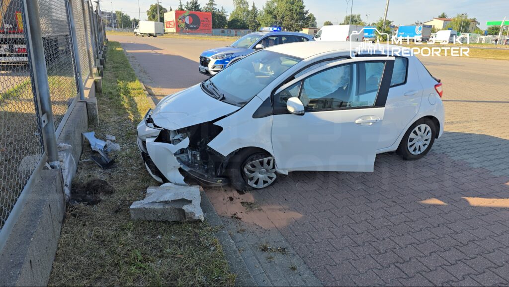 Biały samochód osobowy z uszkodzoną przednią częścią stoi przy ogrodzeniu, obok leżą fragmenty betonu.