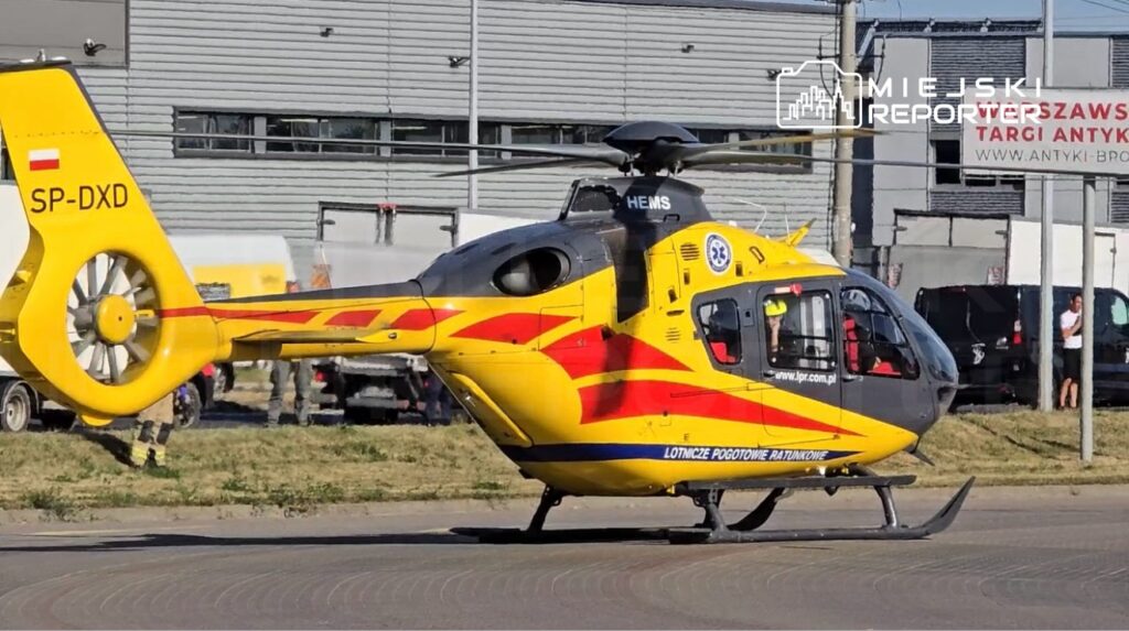 Żółto-czarny śmigłowiec ratunkowy z widocznym napisem "HEMS" i rejestracją SP-DXD stoi na placu przed nowoczesnym budynkiem.
