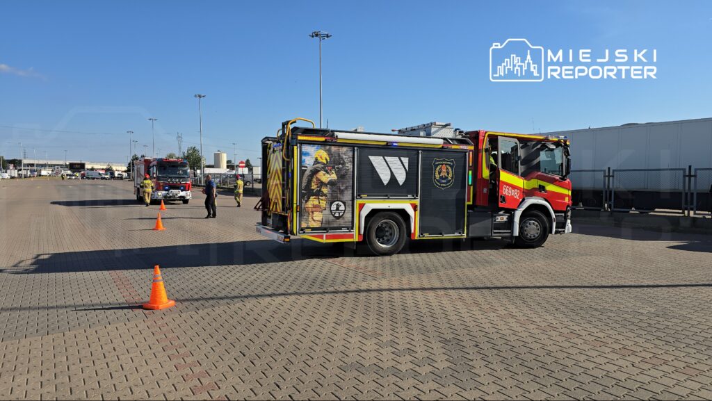 Strażacy w mundurach pracują przy wozie strażackim na terenie parkingu, w tle widoczne inne pojazdy.