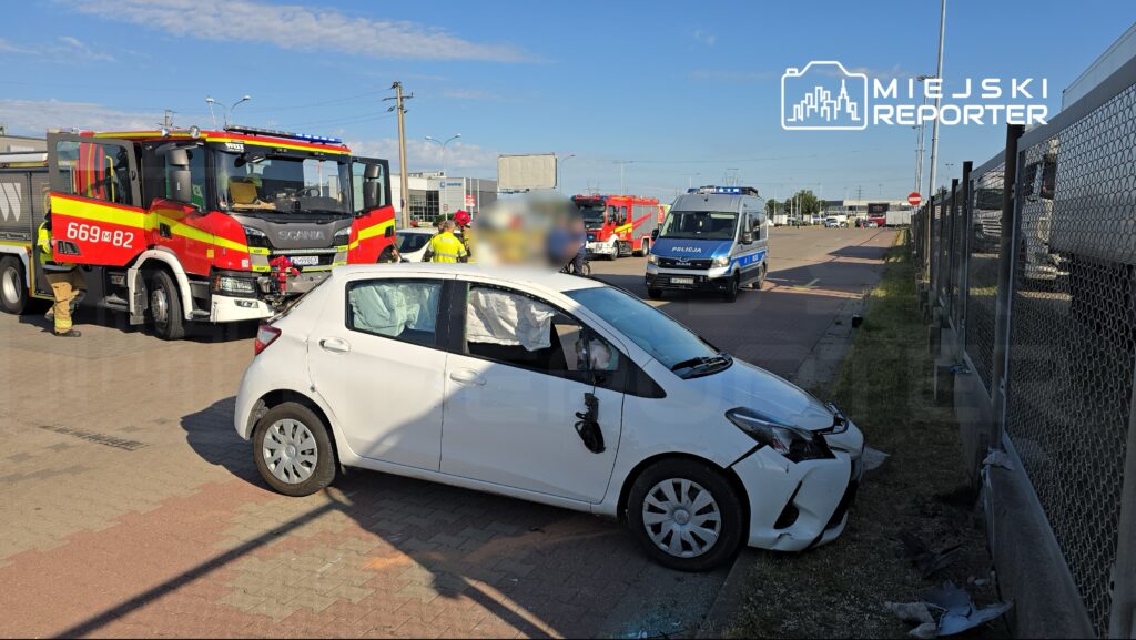 Biały samochód osobowy z uszkodzeniami stoi na poboczu drogi, obok działania służb ratunkowych i policji.