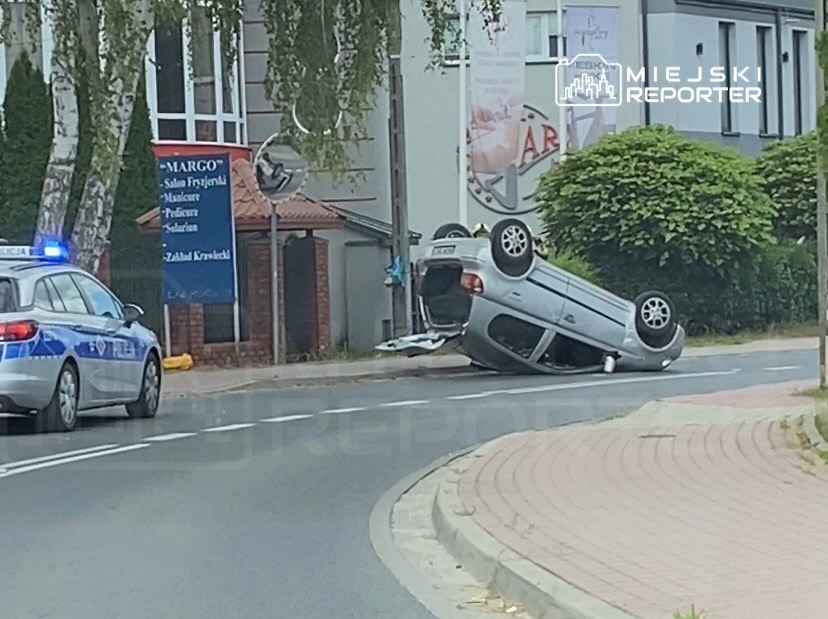 Na drodze leży przewrócone na dach srebrne auto, obok zatrzymany radiowóz z włączonymi sygnałami świetlnymi.