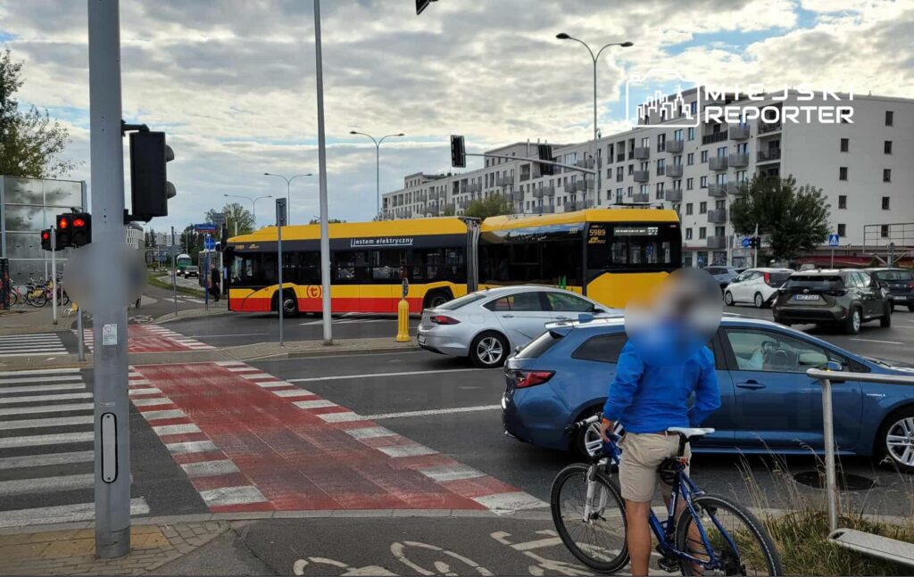 Autobus elektryczny skręca w lewo na ruchliwym skrzyżowaniu, a rowerzysta czeka przy krawężniku.