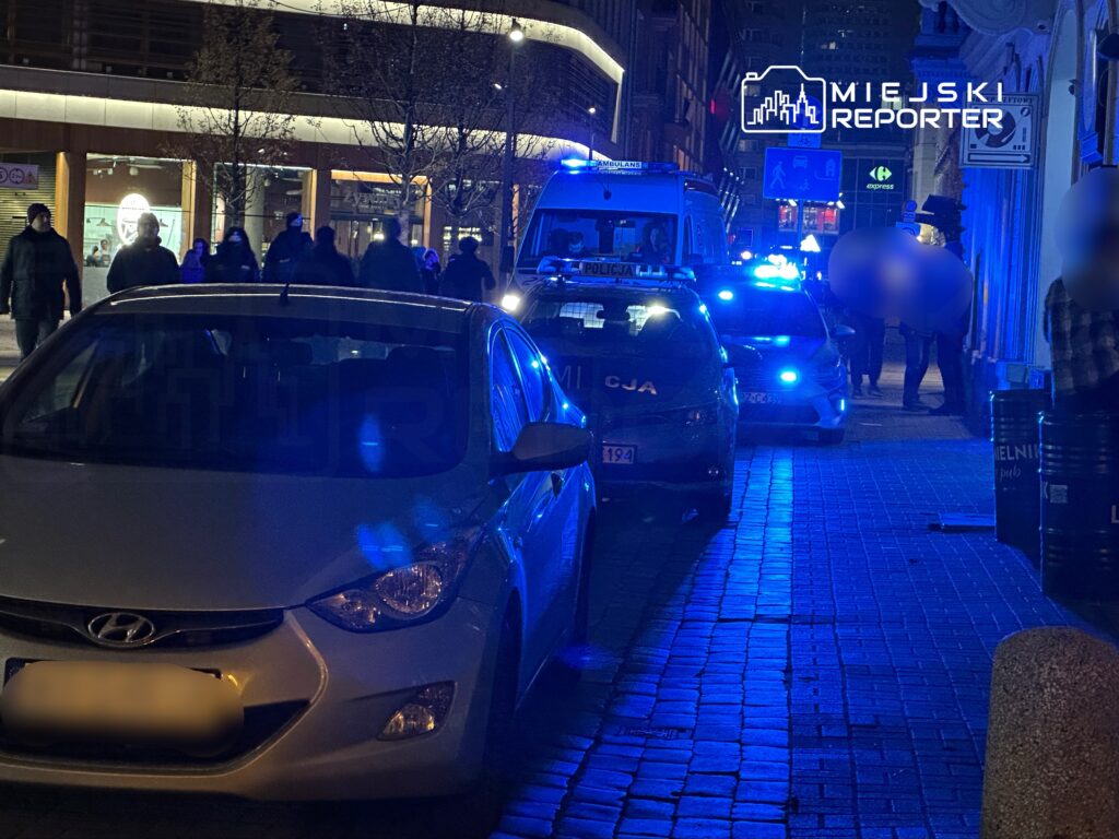 Grupa ludzi spacerujących wzdłuż oświetlonej ulicy, obok zaparkowanych radiowozów policyjnych z włączonymi sygnałami.