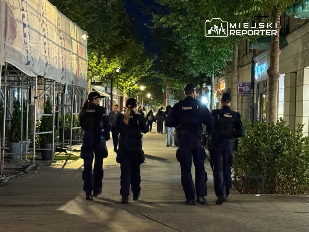 Czterech policjantów patrolujących oświetloną ulicę w nocy, w tle przechodnie i sklepy.