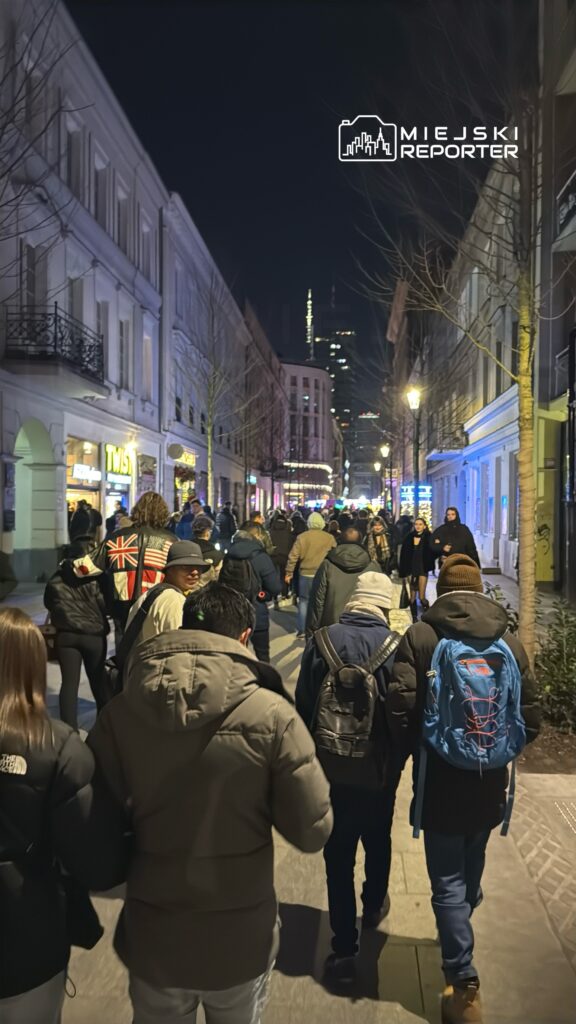 Grupa ludzi spacerująca nocą w oświetlonej ulicy, otoczonej budynkami z witrynami sklepów.