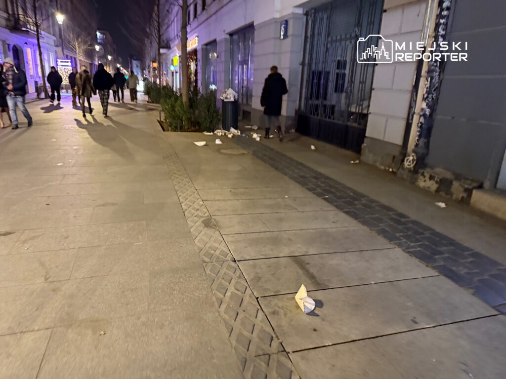 Grupa spacerujących ludzi na oświetlonej ulicy, obok leżące śmieci i zgniecione opakowania.