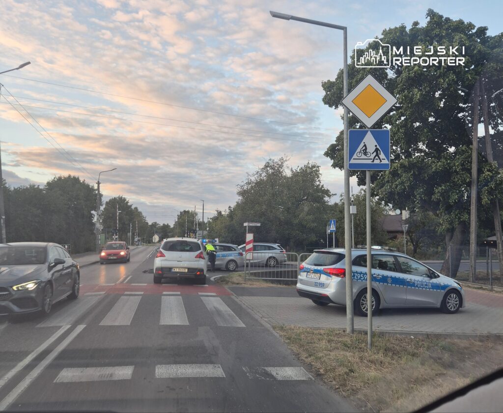 Radiowóz policyjny zatrzymany przy przejściu dla pieszych, obok znaków drogowych, w tle chmurzyste niebo.