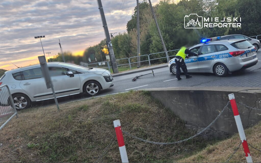 Policjant w kamizelce odblaskowej zabezpiecza miejsce zdarzenia drogowego, obok leży uszkodzona hulajnoga.