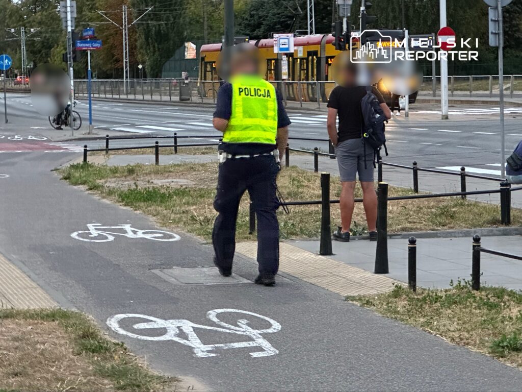 Policjant w kamizelce odblaskowej rozmawia z mężczyzną stojącym obok ścieżki rowerowej w miejskim otoczeniu.