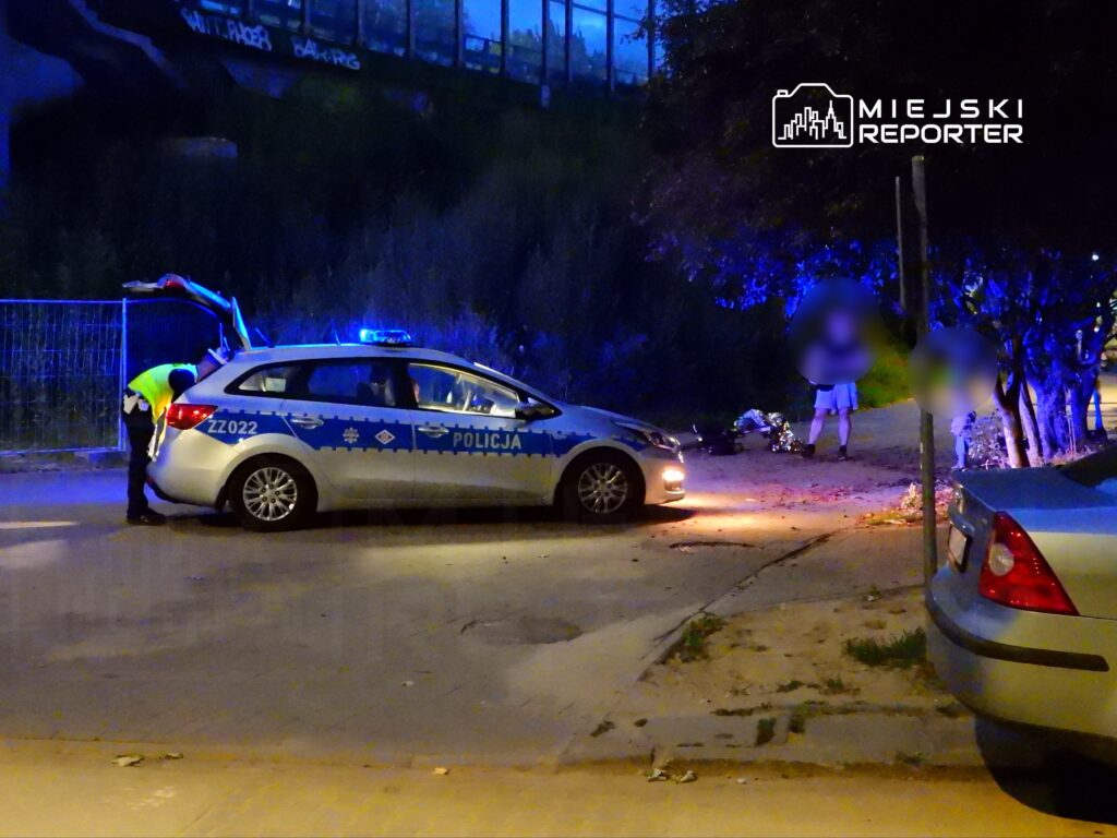 Policjant w kamizelce odblaskowej przeszukuje bagażnik radiowozu na oświetlonej ulicy nocą, obok stoi osoba.