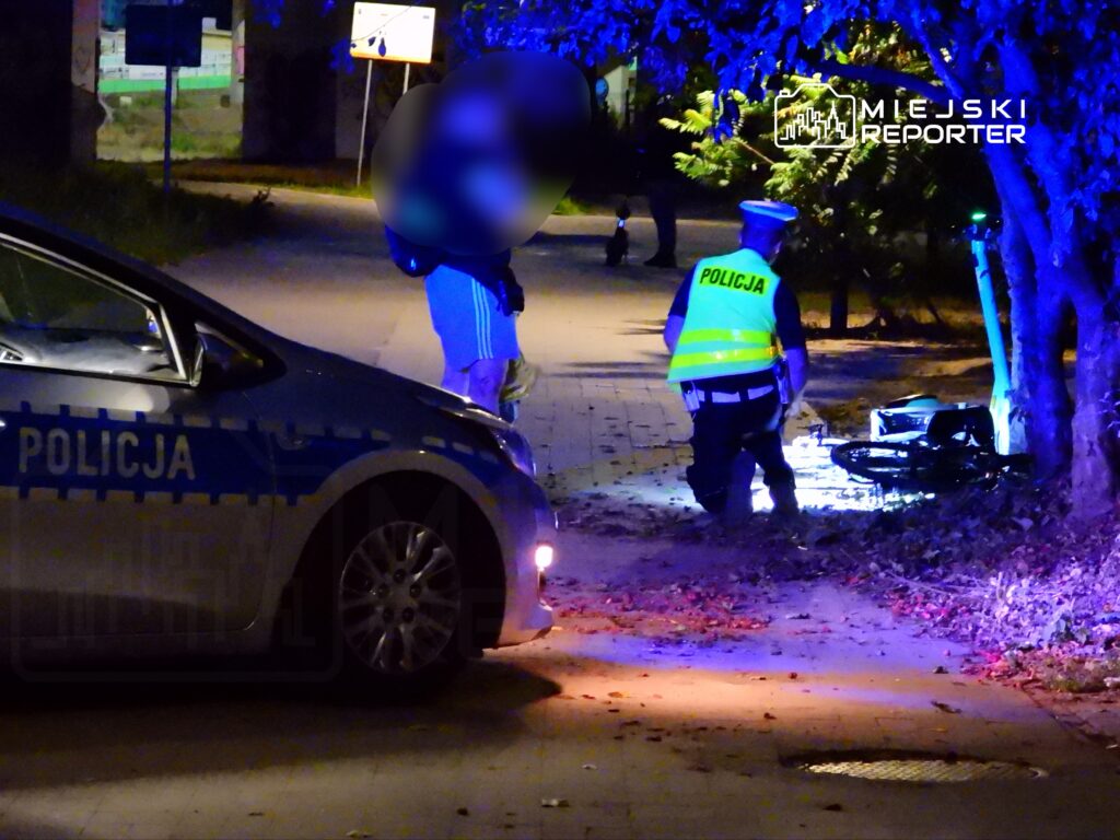 Mundurowy policjant z latarką przeszukuje teren obok przewróconego roweru na oświetlonej ulicy nocą.
