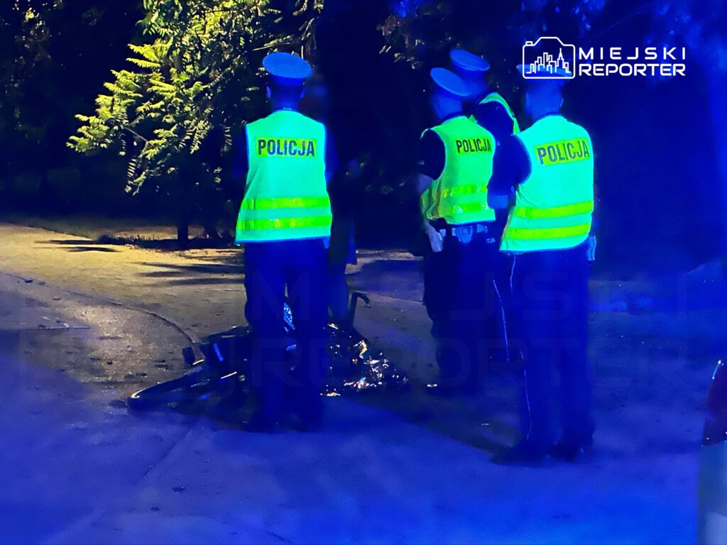Grupa policjantów w kamizelkach odblaskowych prowadzi czynności na oświetlonej ulicy, obok leżącego roweru.