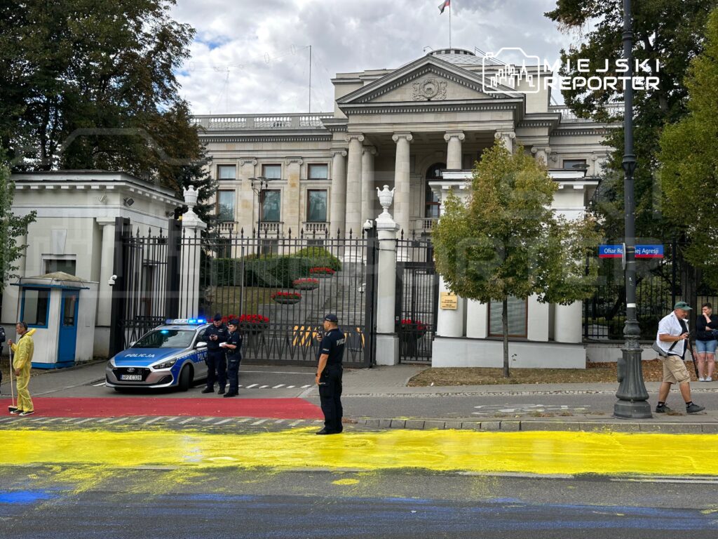 Mężczyzna w żółtym stroju stoi na drodze, obok policyjnego radiowozu, na tle monumentalnego budynku z kolumnami.
