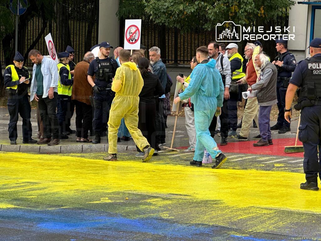 Grupa ludzi, w tym policjanci, uczestniczy w wydarzeniu na ulicy, gdzie na jezdni leży żółty proszek.