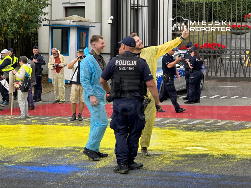 Grupa osób w strojach ochronnych maluje nawierzchnię na żółto, podczas gdy policjanci monitorują sytuację.