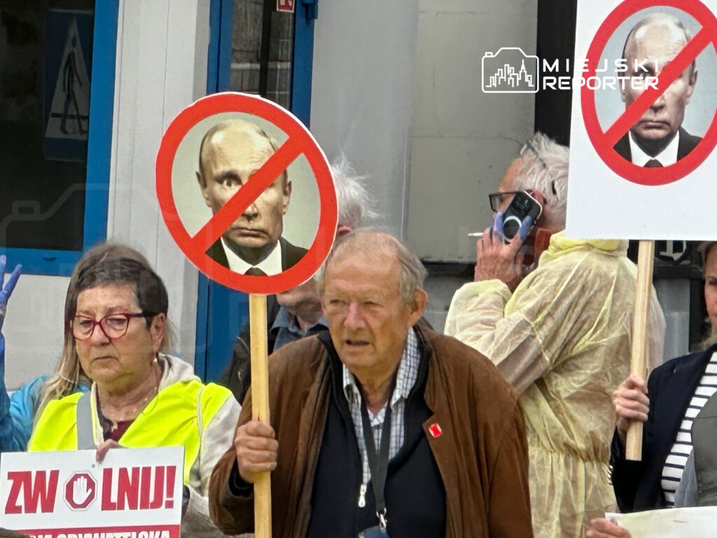 Grupa protestujących z transparentami i plakatami z przekreślonym wizerunkiem mężczyzny w garniturze, stojąca na ulicy.