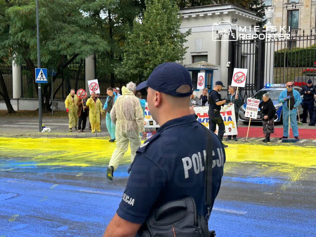 Grupa protestujących w kombinezonach ochronnych maluje jezdnię na żółto i niebiesko, trzymając transparenty. Policjant obserwuje sytuację.