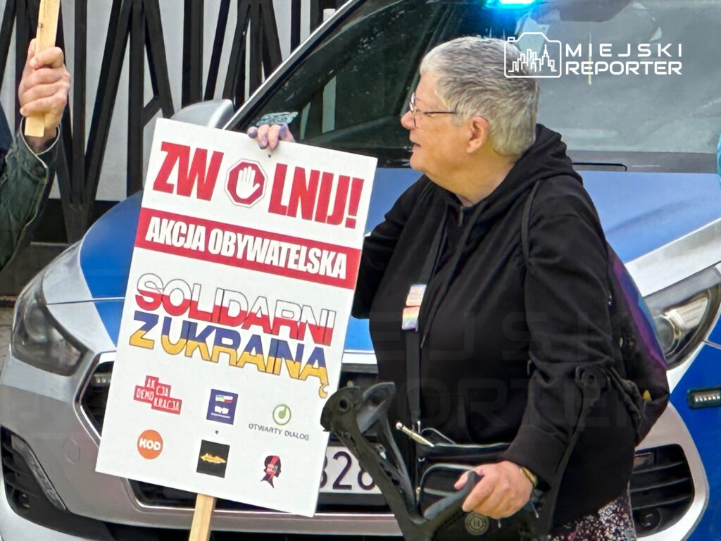 Kobieta w okularach trzyma transparent z napisem "ZWOLNIJ! AKCJA OBYWATELSKA SOLIDARNI ZUKRANIA" obok radiowozu.