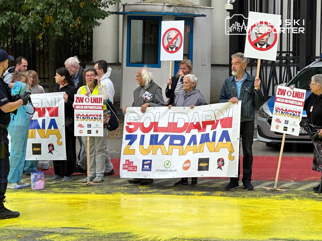 Grupa osób z transparentami i plakatami protestuje na ulicy, w tle widoczny radiowóz oraz żółta farba na jezdni.
