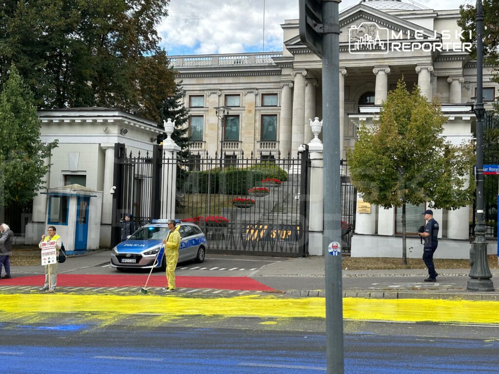 Dwie osoby malują kolorowe pasy na jezdni przed budynkiem z kolumnami, obok stoi radiowóz policyjny.