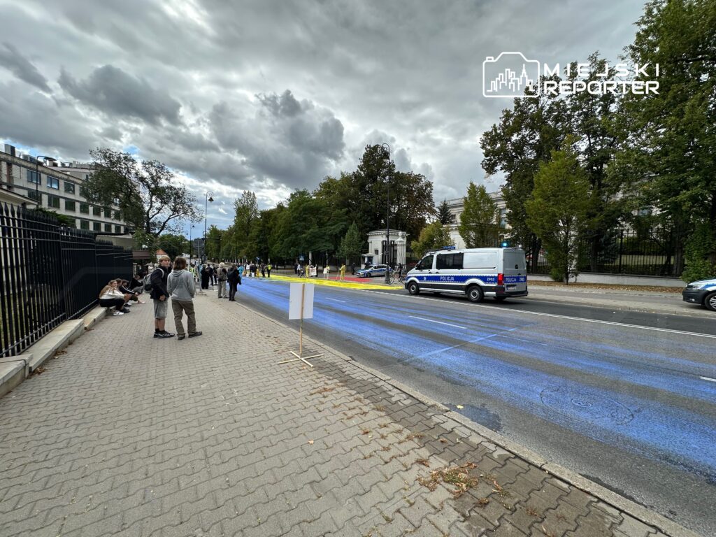 Grupa ludzi stoi na chodniku obok ulicy, gdzie policyjny radiowóz przejeżdża obok niebieskiej farby na jezdni.