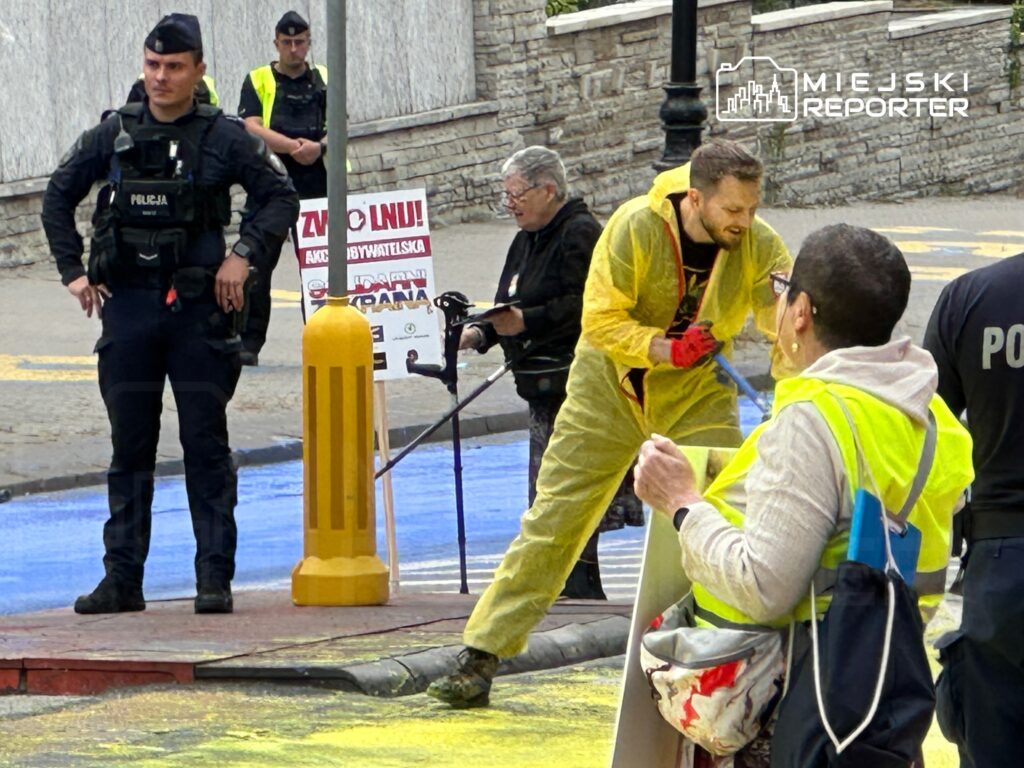 Mężczyzna w żółtym kombinezonie maluje chodnik, a obok niego stoi policjant i kilka osób w odblaskowych kamizelkach.