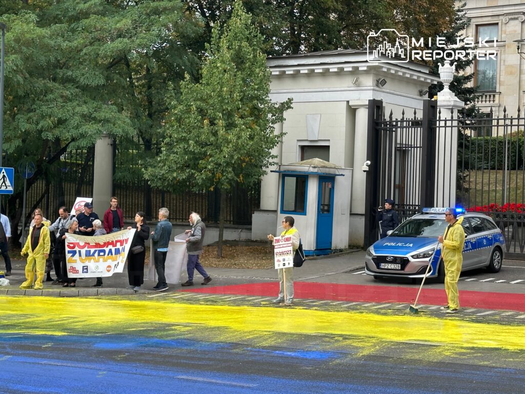 Grupa osób trzymająca transparent "Solidarni z Ukrainą" protestuje, a w tle mężczyzna w żółtym kombinezonie maluje drogę na kolorowo.
