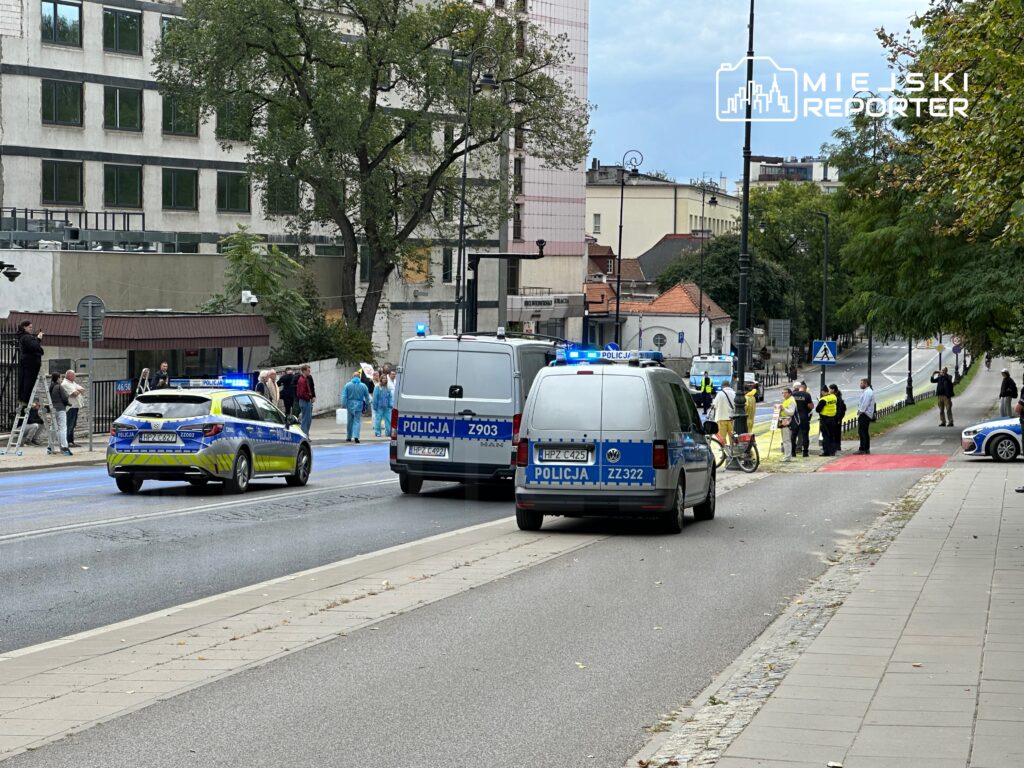 Grupa policjantów zabezpiecza teren przy drodze, gdzie stoją radiowozy i zbiera się tłum ludzi.