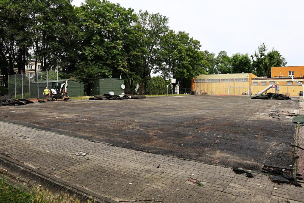 Pracownik w żółtej kamizelce obsługuje małą maszynę budowlaną na opuszczonym boisku, wokół leżą resztki materiałów.