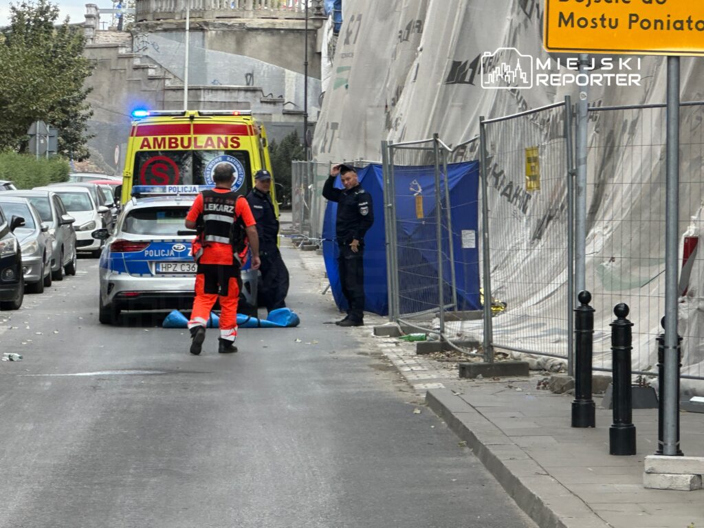 Ratownik medyczny w pomarańczowym uniformie podchodzi do policjantów przy zaparkowanym radiowozie na ulicy z budową w tle.