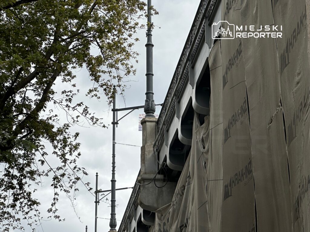 Fragment budynku pokryty materiałem ochronnym, z widocznymi elementami architektonicznymi i gałęziami drzew w tle.