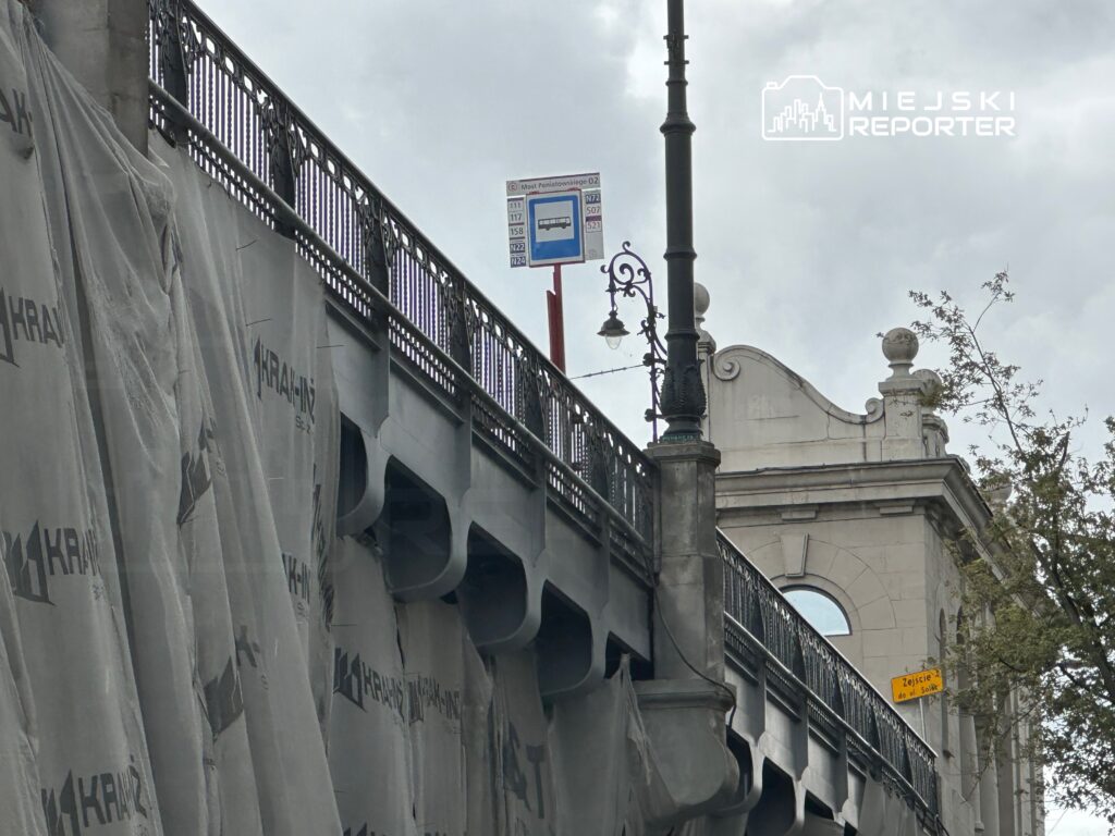 Most z przeszkloną balustradą, zasłonięty siatką budowlaną, z oznaczeniem przystanku autobusowego w tle.