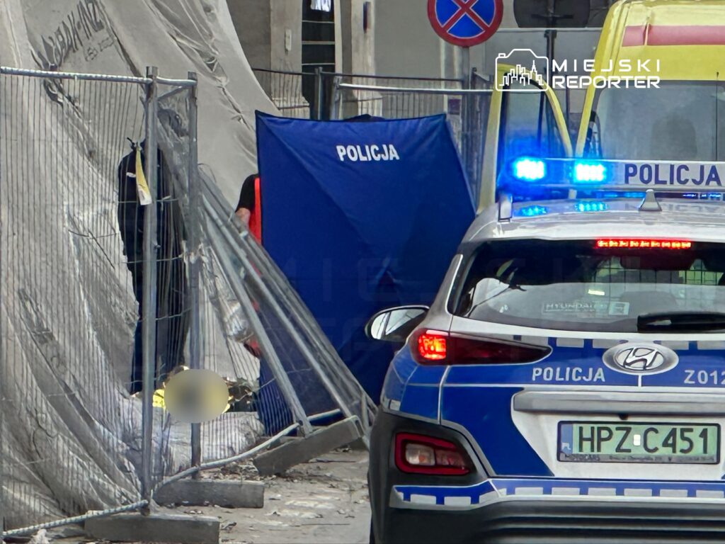 Funkcjonariusze policji przy zasłonie na miejscu zdarzenia, obok radiowozu z włączonymi sygnałami świetlnymi.