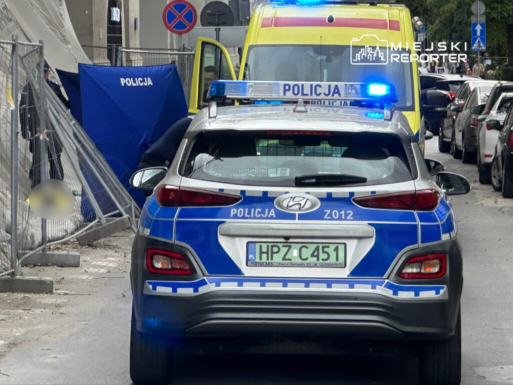 Policyjny radiowóz z niebieskimi światłami zaparkowany na ulicy obok karetki, w tle materiał policyjny.