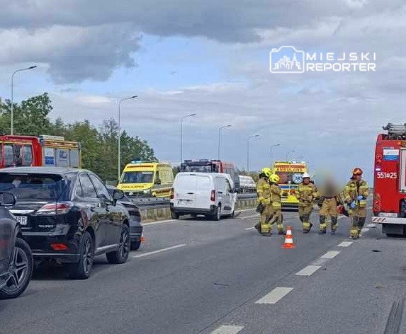 Grupa strażaków w mundurach przeprowadza działania ratunkowe na drodze, obok stojących pojazdów służb ratunkowych.