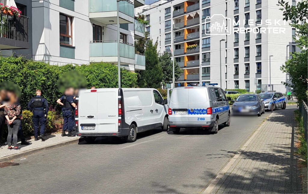 Grupa policjantów rozmawia z dwiema osobami na chodniku, obok zaparkowanych radiowozów i furgonetek.