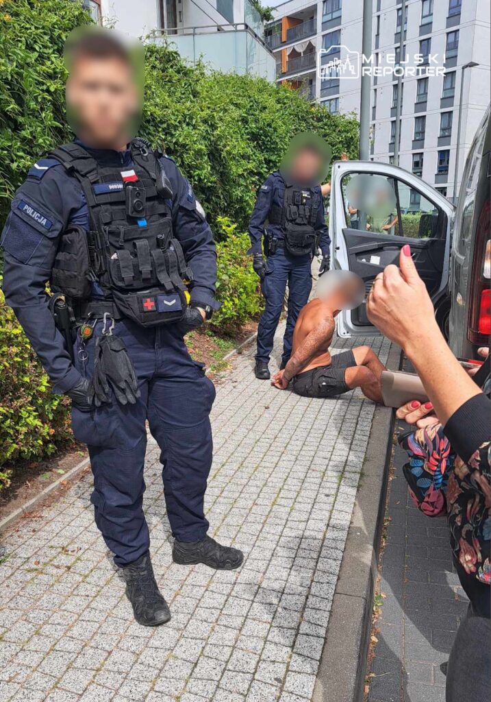 Dwaj policjanci w mundurach stoją obok zatrzymanego mężczyzny na chodniku, a kobieta gestykuluje w jego kierunku.