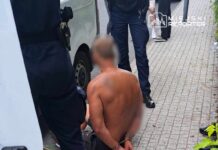 Mężczyzna w kajdankach siedzący na chodniku, otoczony przez policjantów w umundurowaniu.